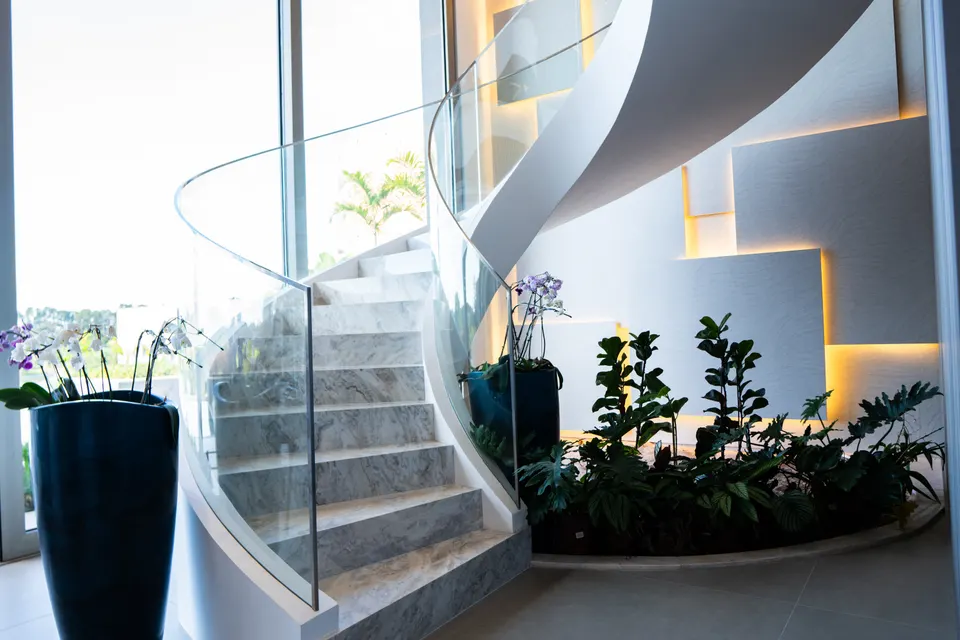 Escada curva com guarda-corpo de vidro, jardim interno e iluminação indireta